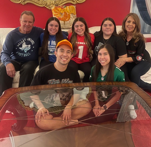 Group of seven people (two adults, five teens) sitting on a couch in a red-walled living room, smiling at the camera around a glass coffee table.