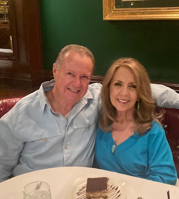 Smiling man in a light blue shirt and woman in a turquoise top sit close together at a restaurant table, with a chocolate dessert on a plate in front of them.