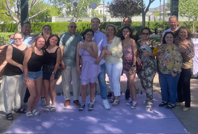 Don Salazar and his large family posing for a group photo on a lavender carpet outdoors on a sunny day, some holding flowers, everyone smiling at the camera.