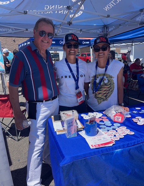 Three people pose at an outdoor booth under a blue canopy; two women wear 76 hats and lanyards, a man in a striped shirt stands with them.