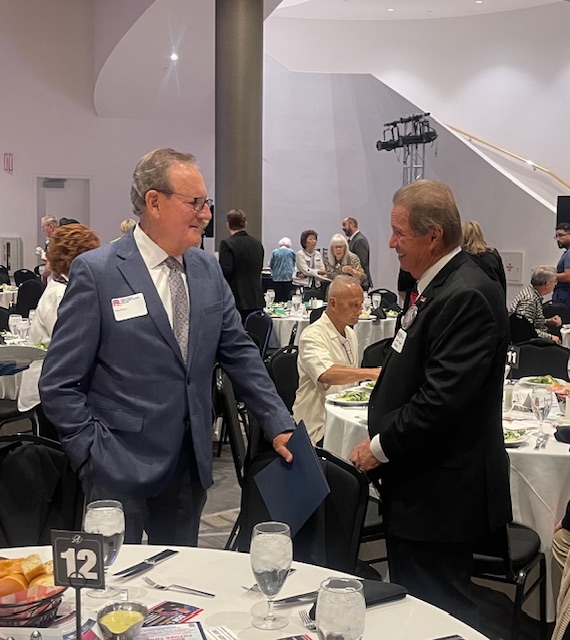 Two men in suits shake hands at a round-table banquet during a formal event.