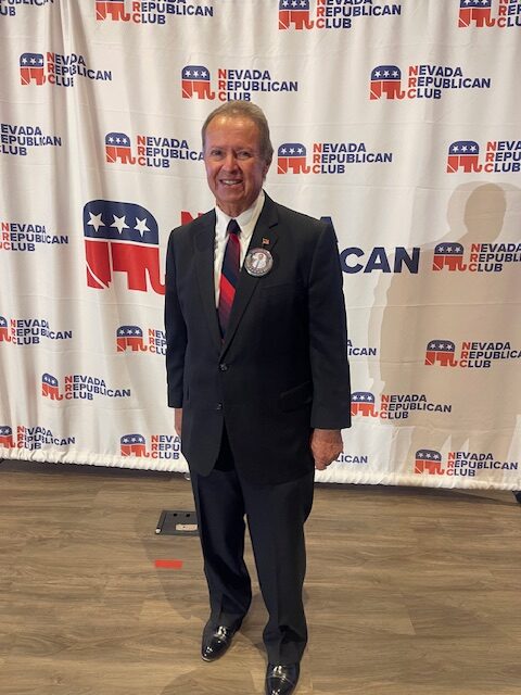 Older man in a dark suit and red tie standing in front of a Nevada Republican Club backdrop with a circular badge on his lapel.