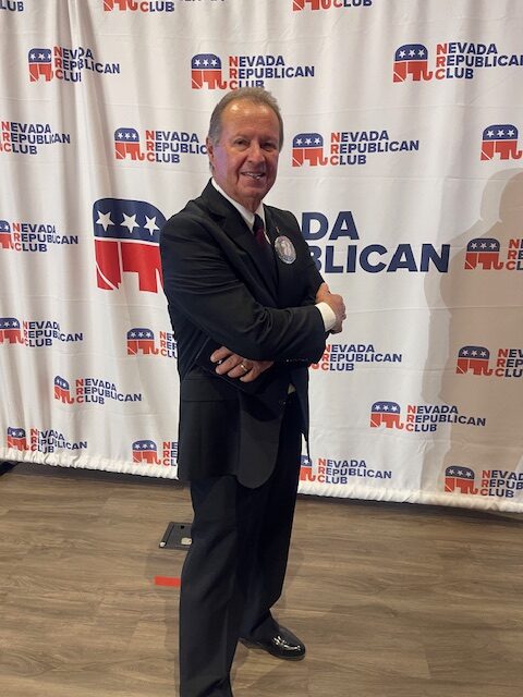 Man in a dark suit with a red tie stands with arms crossed in front of a Nevada Republican Club backdrop.