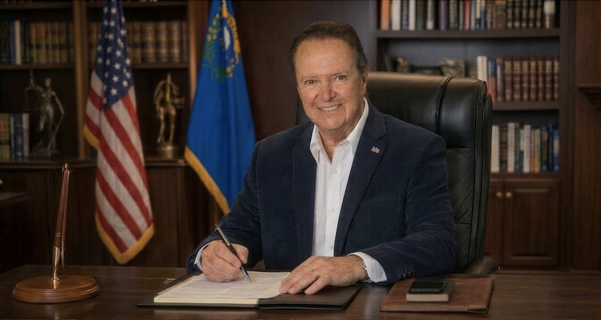 Smiling man in a dark blazer signs a document at a wooden desk in a formal office, with the American flag and state flag in the background.