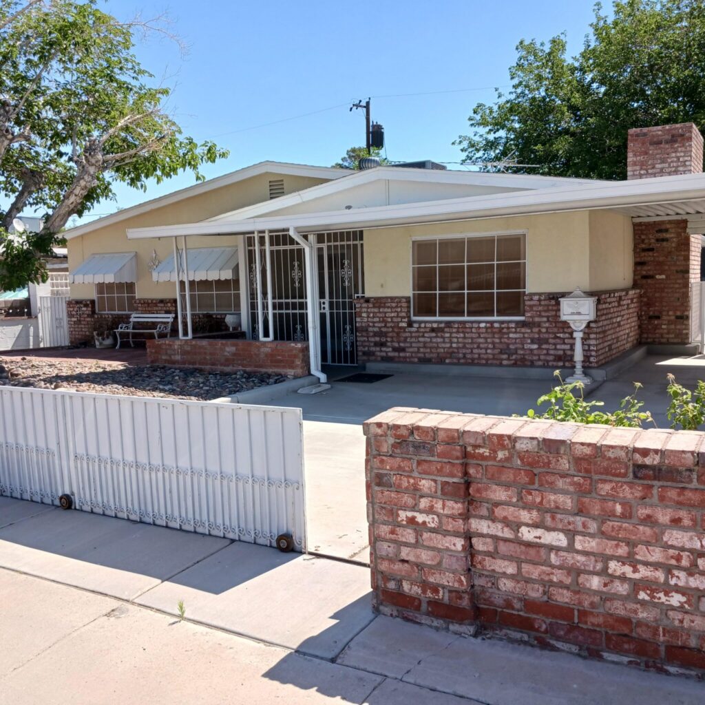 Front view of a single-story beige house with brick accents, white gate, and a paved driveway under a clear blue sky.