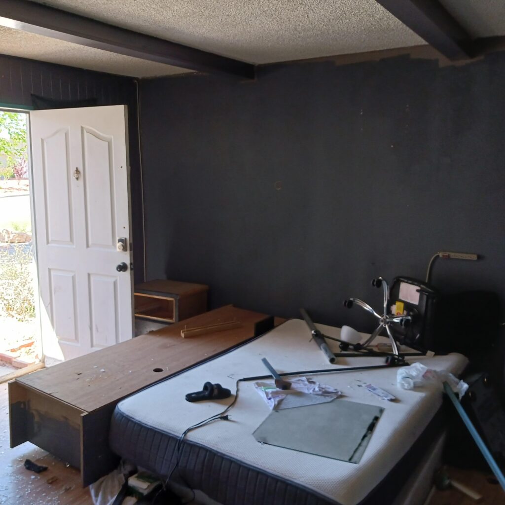 Dimly lit room with dark walls and exposed ceiling beams, an open white door, and a partially disassembled sofa bed with clutter on the floor outside the door edge.
