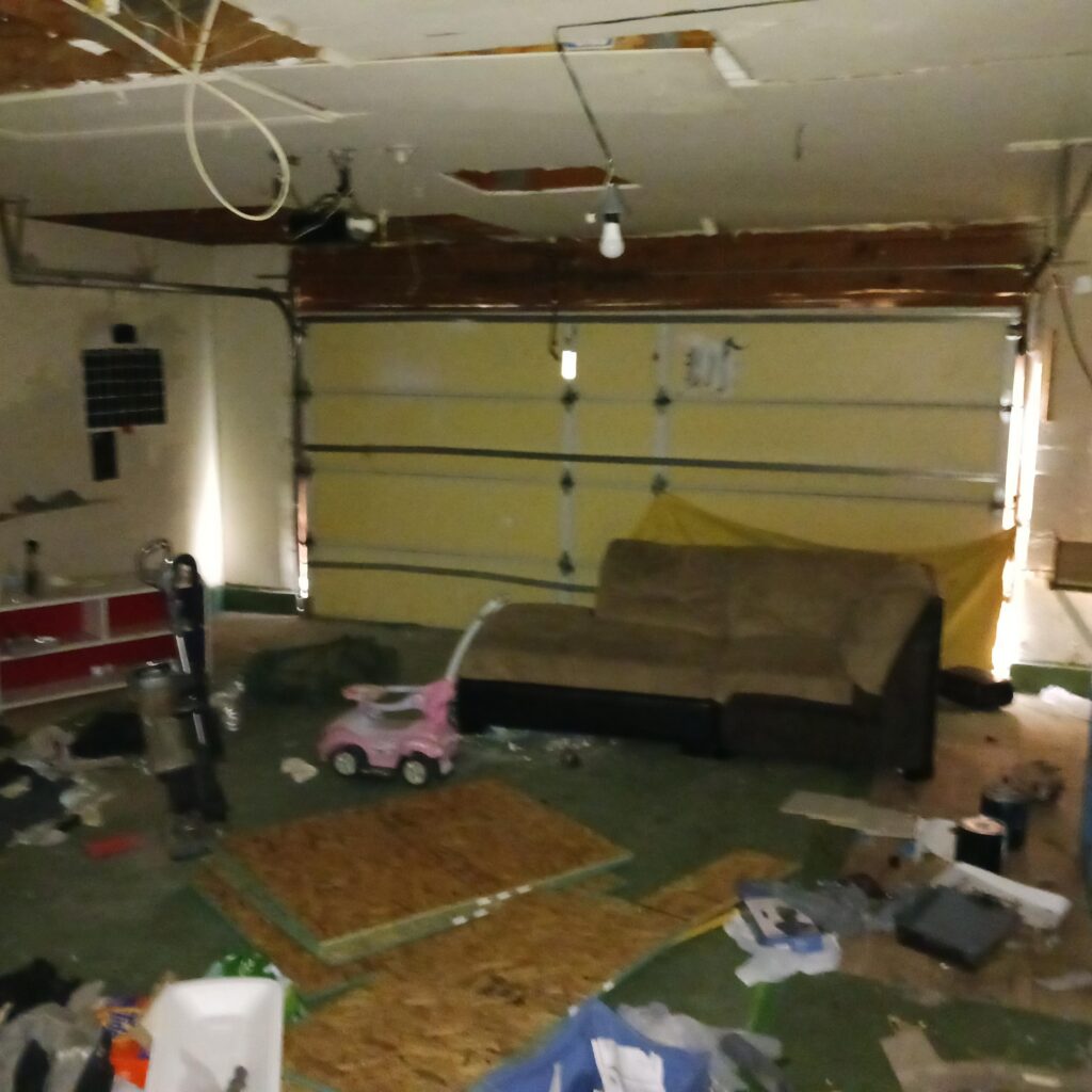 Messy garage or basement scene with scattered toys, papers, and clutter on the floor and shelves; exposed ceiling and walls showing wear.