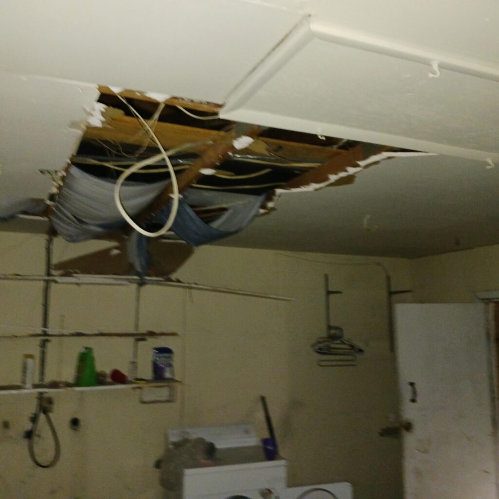 Basement utility room with a damaged ceiling, exposed beams and wiring above a wringing mess of laundry items and clutter on the floor.]