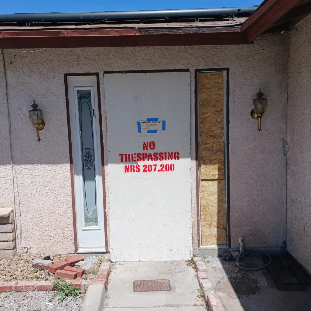 Front porch of a stucco house with a boarded-up entry door and a 'No Trespassing' sign taped to the board; construction debris on the ground.