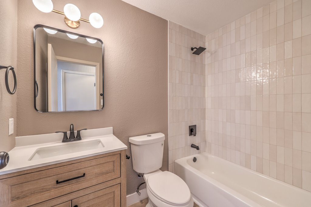 Bathroom with a wooden vanity, white countertop and sink, black faucet, and a large rectangular mirror above; a toilet beside the vanity and a bathtub with white tiled walls to the right; warm lighting above.