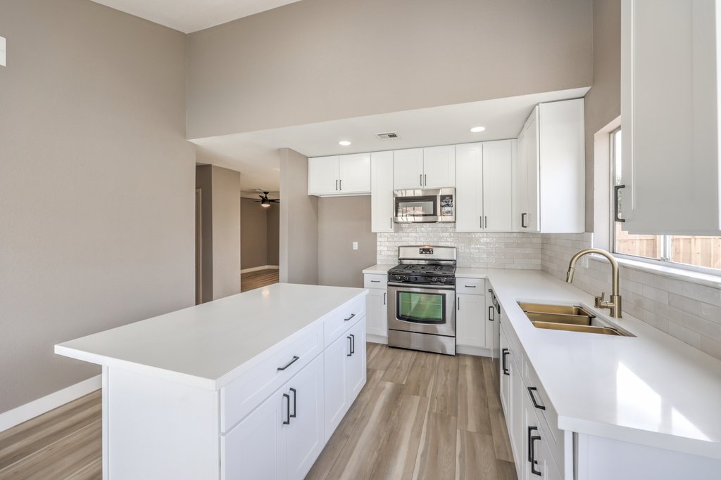 Bright white modern kitchen with an island, white cabinets, stainless-steel appliances, and a double sink under a large window.