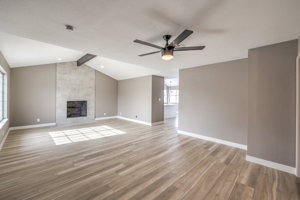 Spacious open-concept living room with gray walls, light wood floors, a modern fireplace, and a ceiling fan, bathed in natural light from large windows on the left.